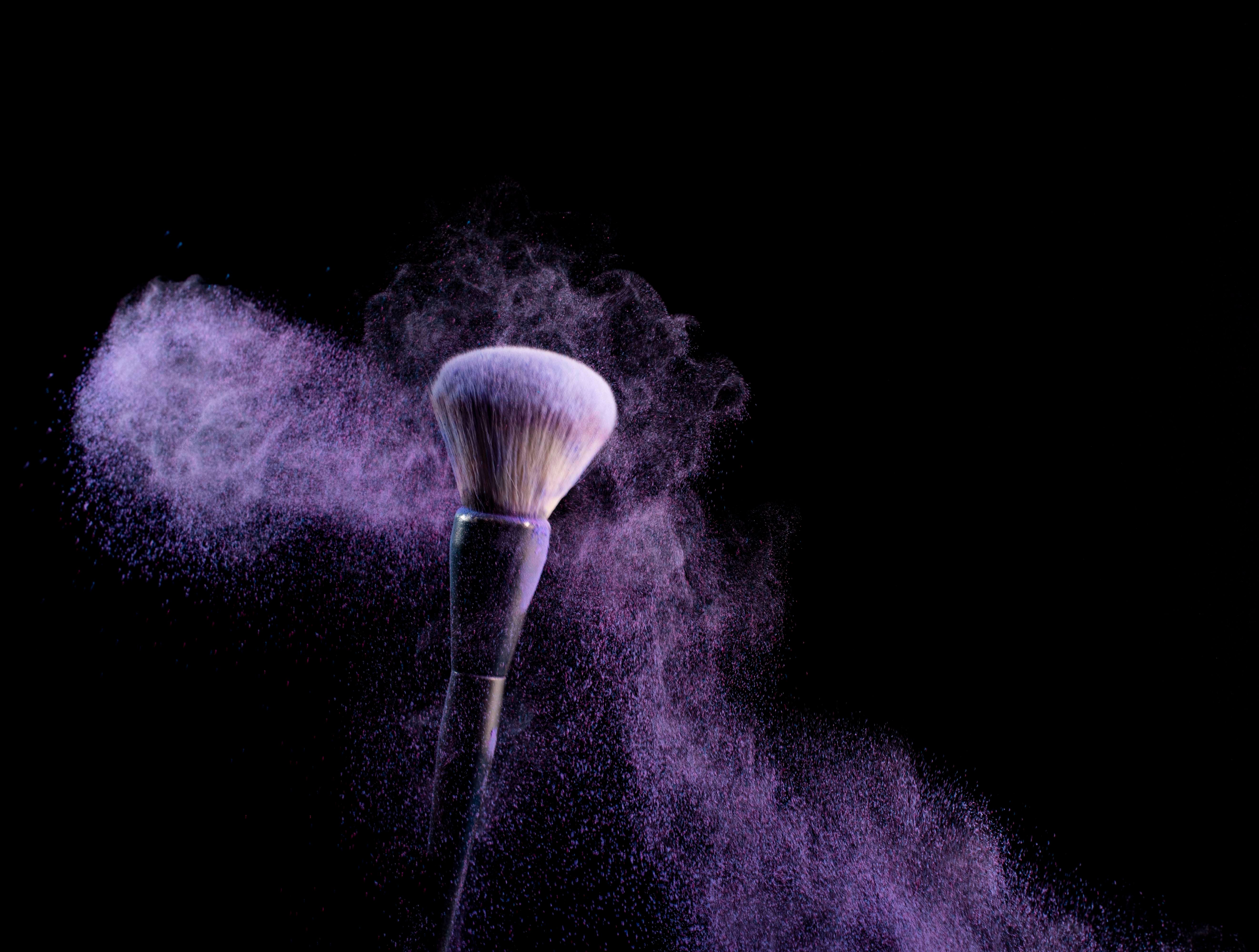 Fotomural Pincel de maquillaje con explosión de polvo morado sobre fondo negro, resaltando la belleza y el arte del maquillaje.