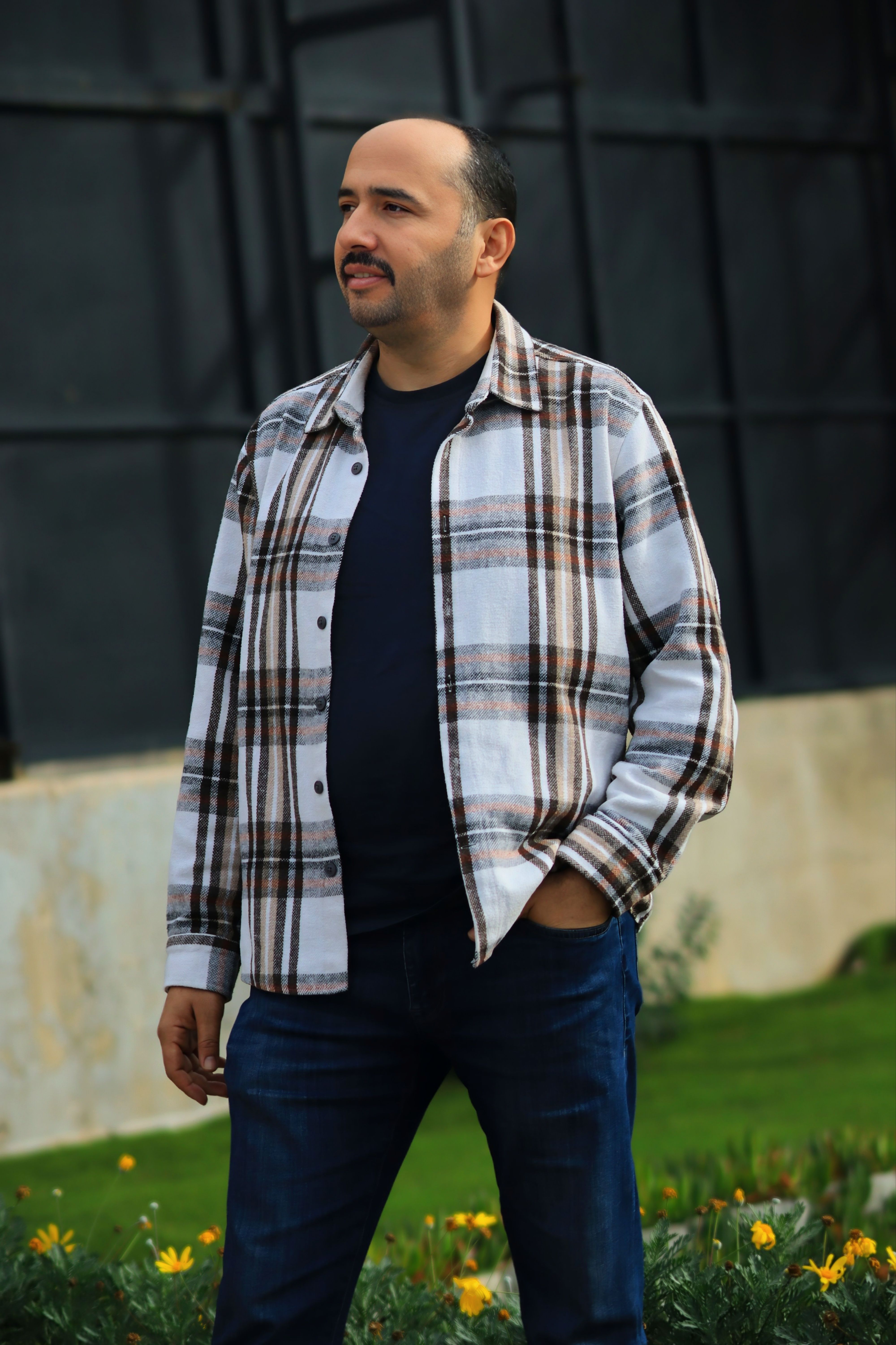 A man wearing a plaid shirt and jeans standing in a garden setting.