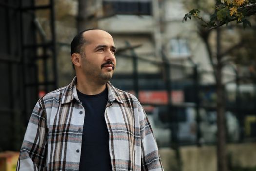 A man in plaid shirt outdoors, looking thoughtful, against an urban backdrop. Captured in fall.