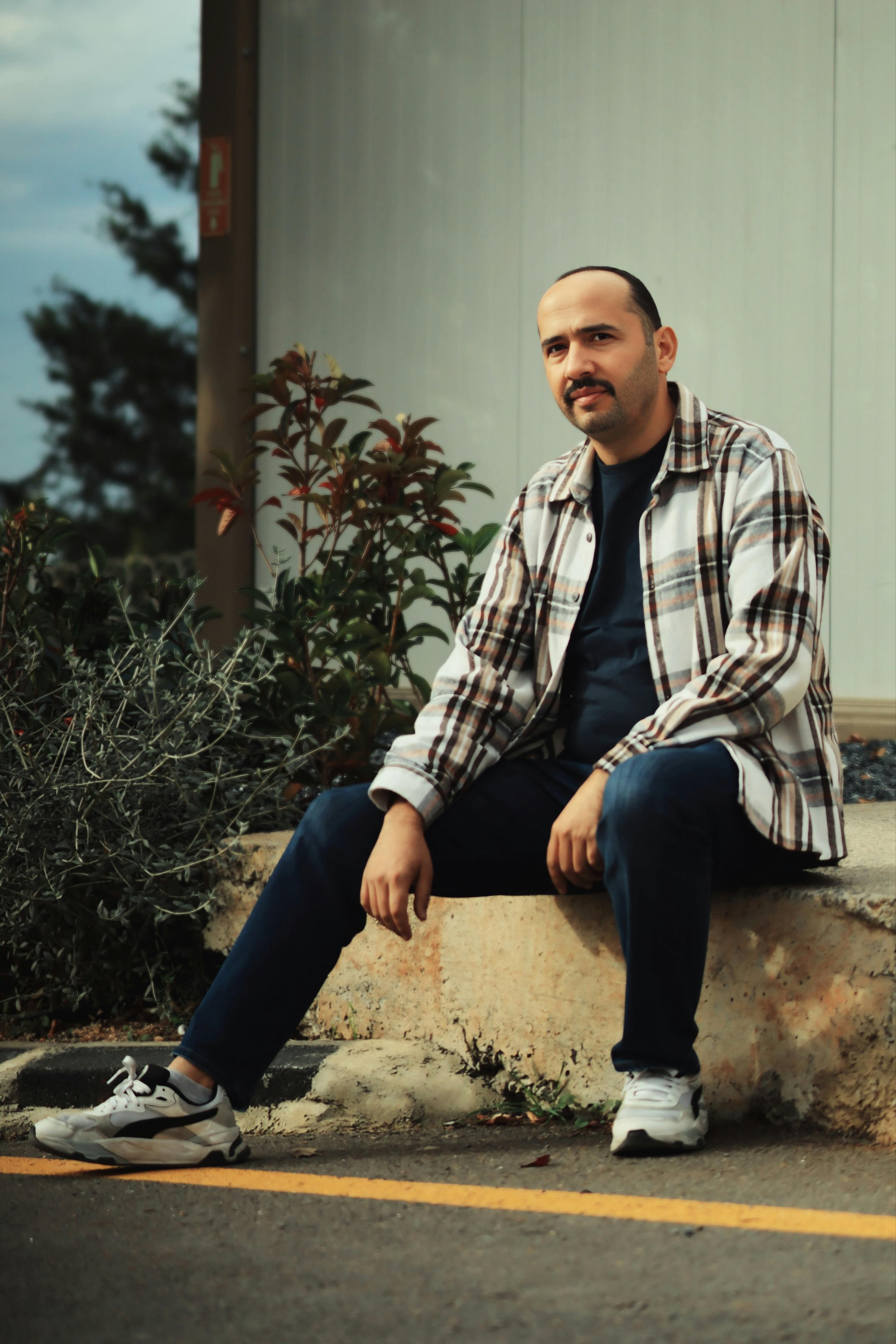 Man sitting outdoors in casual attire, wearing a plaid shirt, jeans, and sneakers, with a relaxed demeanor.