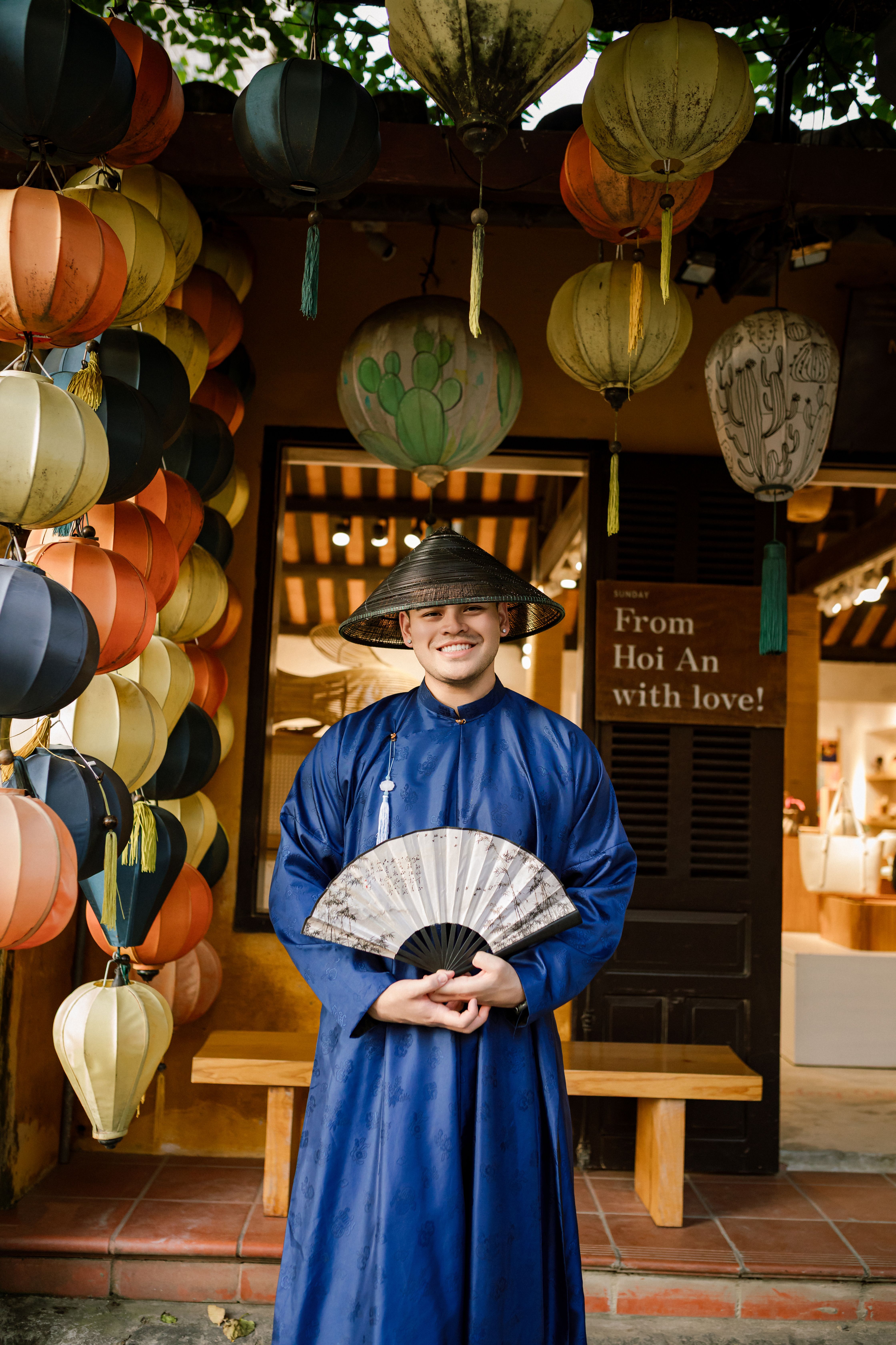 Man in traditional blue attire with a fan, standing in a Hội An lantern shop.