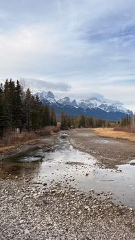 Tranquil river flowing through rocky landscape with snow-capped mountains in the background under a cloudy sky.