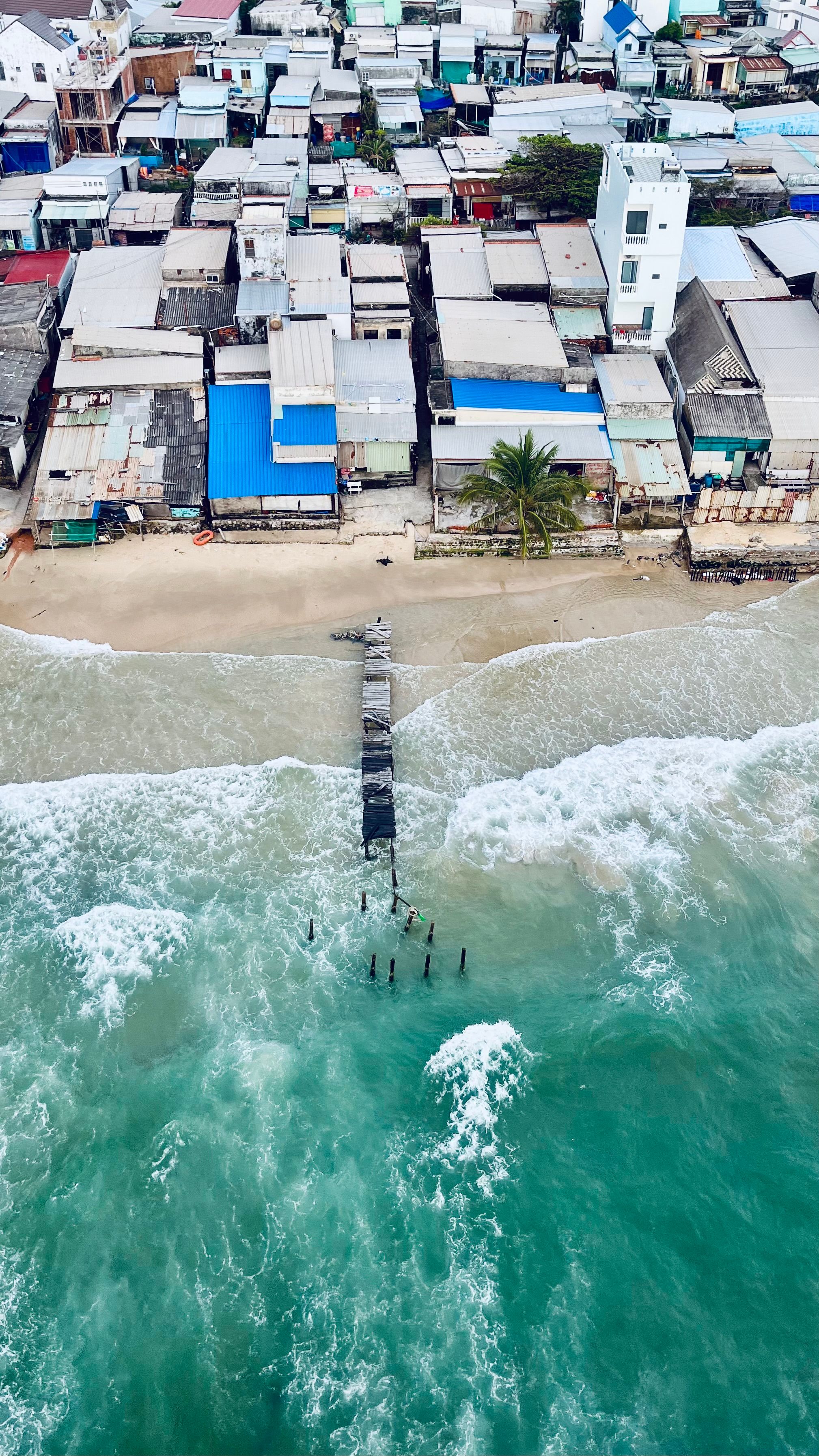 Aerial shot of a coastal town with diverse rooftops and a sandy beach meeting the turquoise sea.