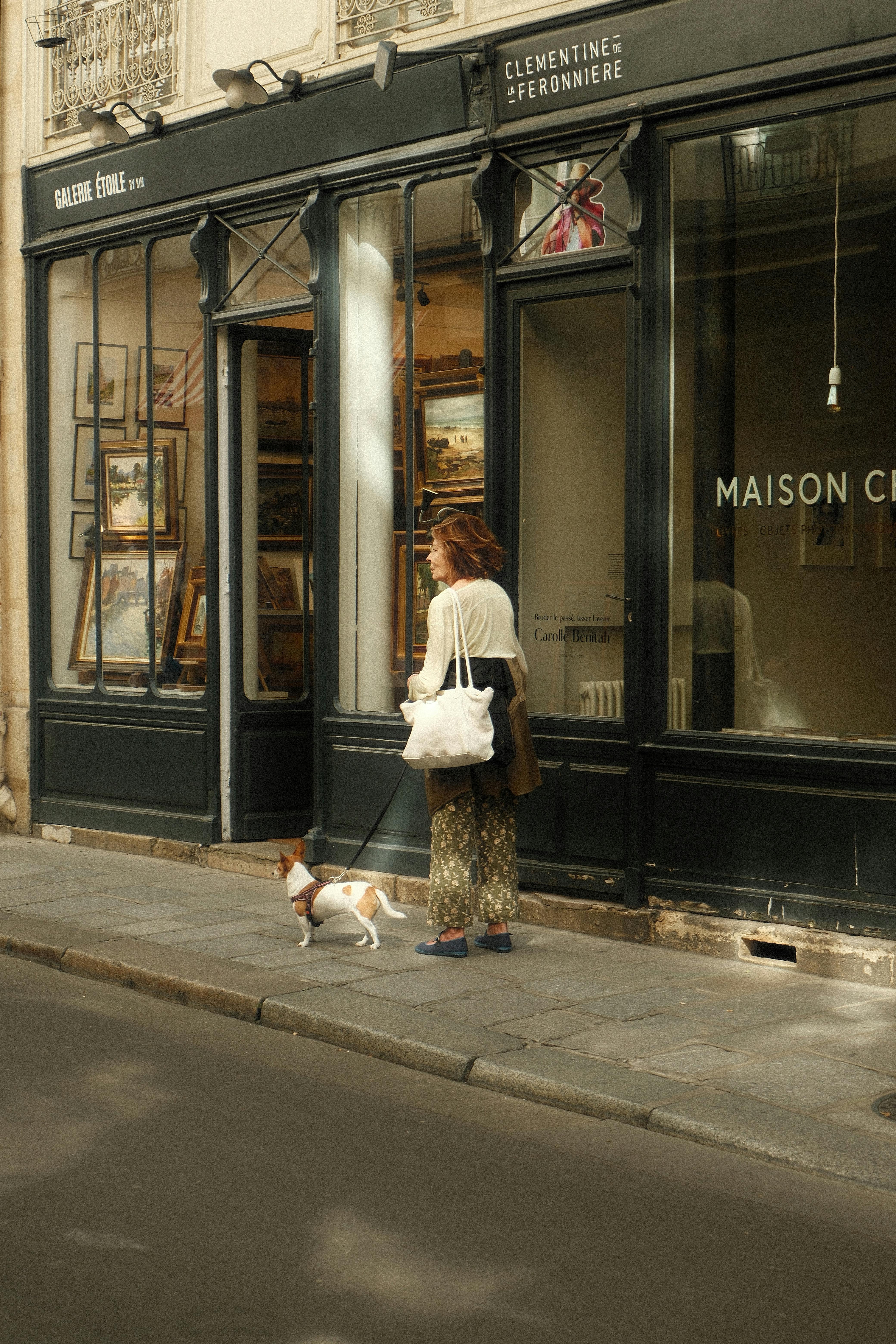 A woman walks her dog past charming Parisian shops on a sunny day.