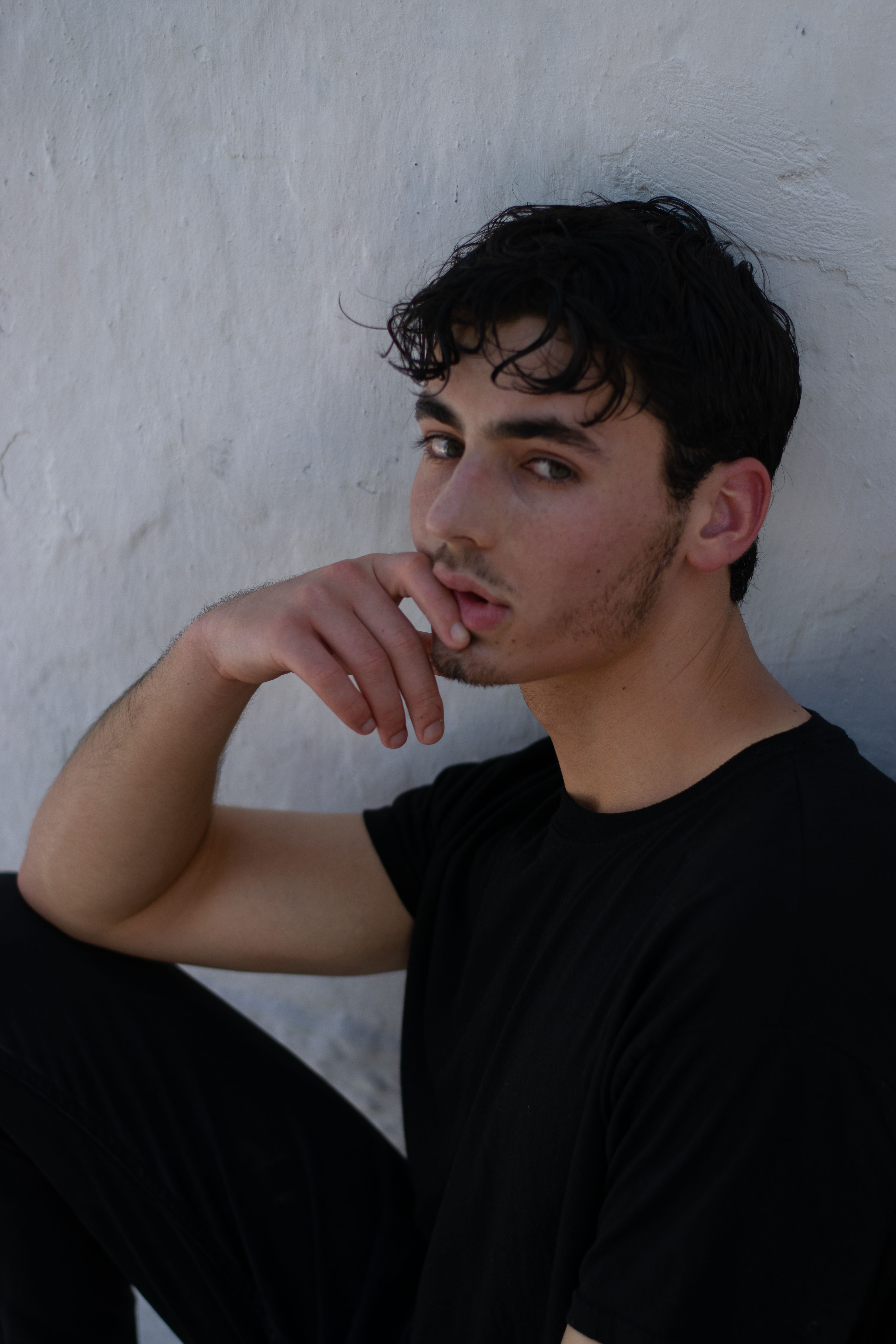 Profile portrait of a young man in black clothing, looking thoughtful against a white wall.