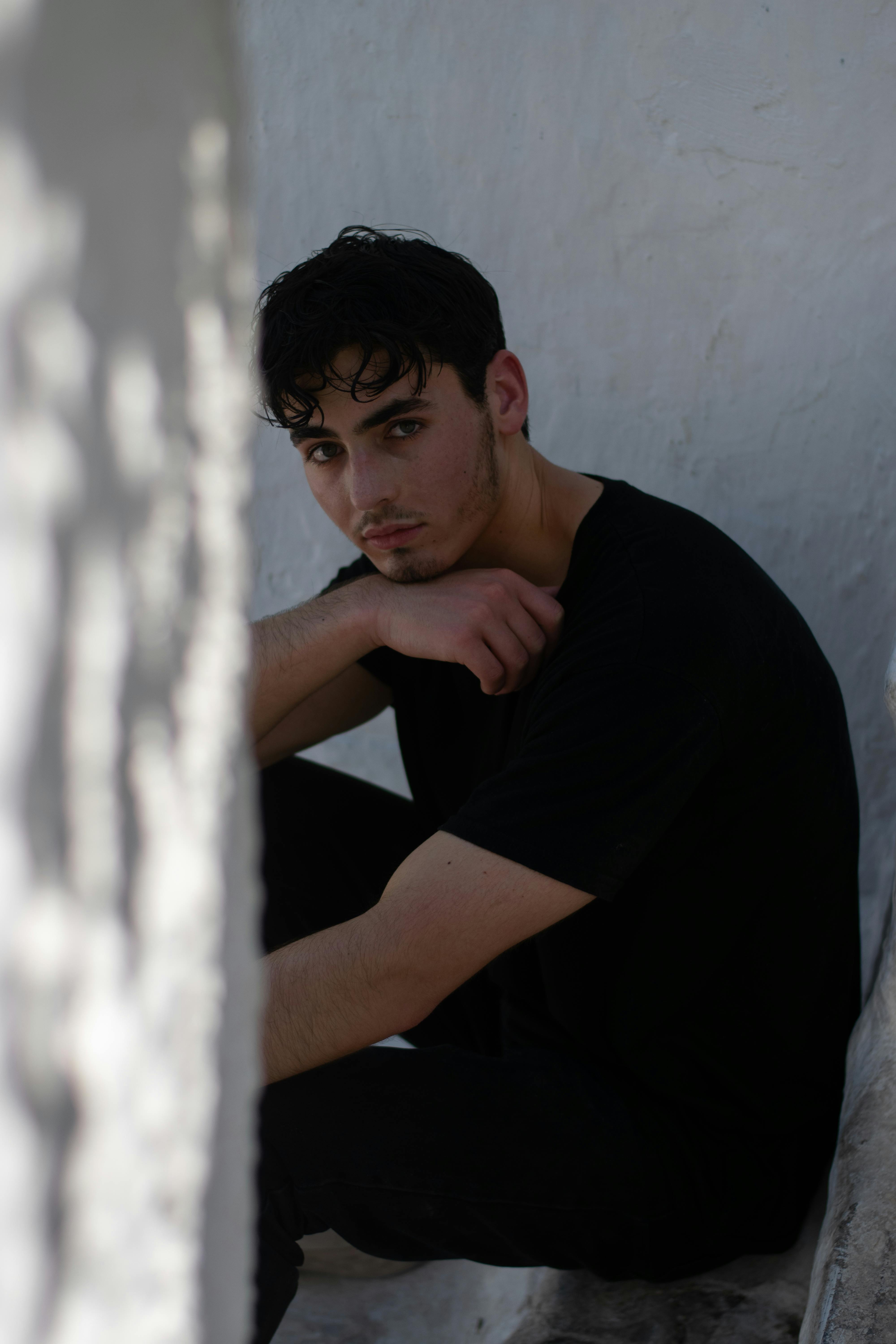 Young man sitting indoors in low light, conveying a thoughtful, introspective mood.