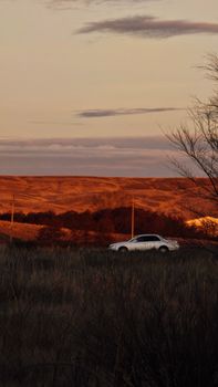 A lone white car on a rural landscape during sunset with golden hills in the background.