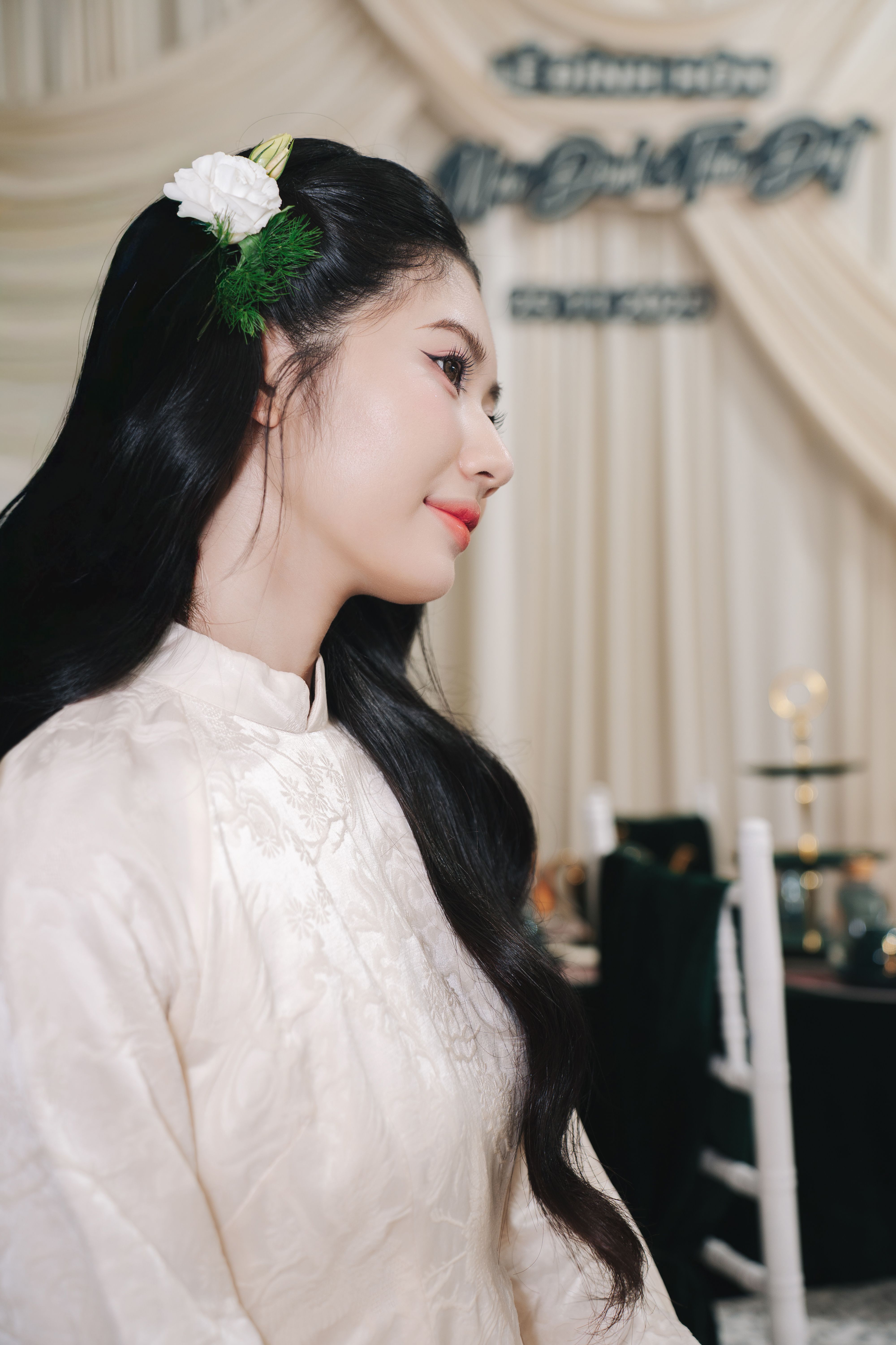 Side profile of a woman in a white traditional dress with floral hair accessory at a formal gathering.