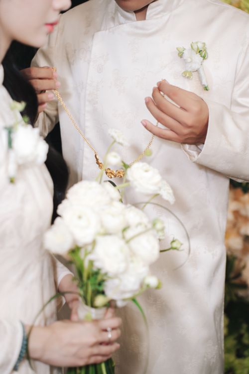 Free Close-up view of a couple in white wedding attire exchanging a gold necklace with white flowers. Stock Photo