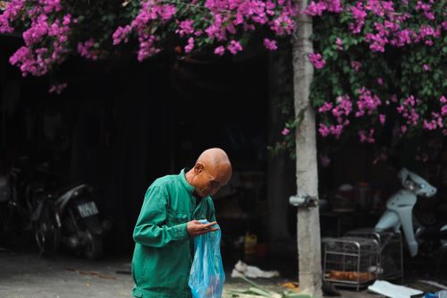 Free Elderly man stands beneath vibrant pink bougainvillea in a busy urban street. Stock Photo