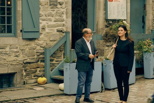 Two people conversing outside a stone building in Quebec, showcasing historical architecture and autumn decor.