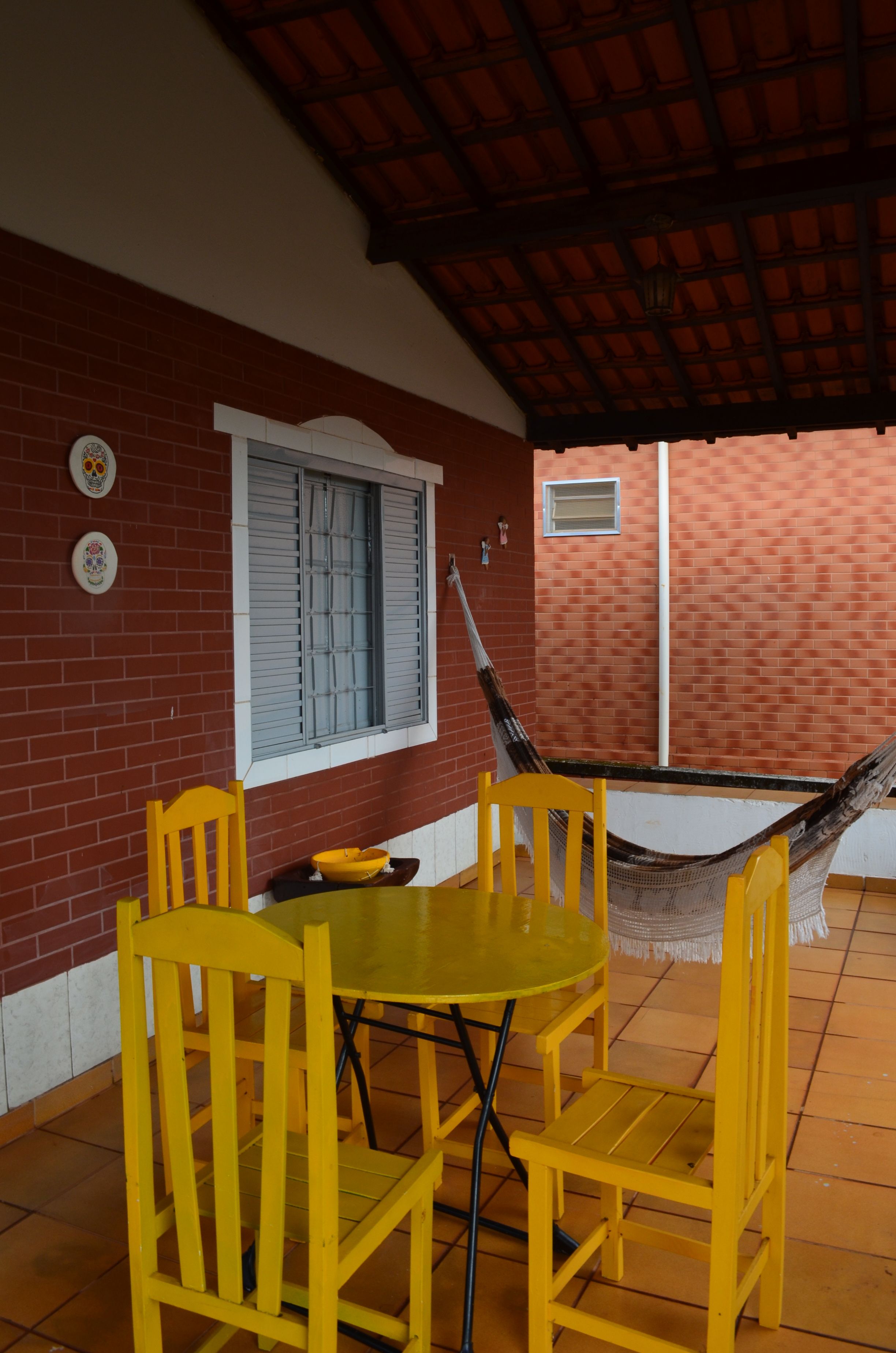 Mesa e cadeiras de madeira amarelas numa varanda de tijolo, com rede ao fundo