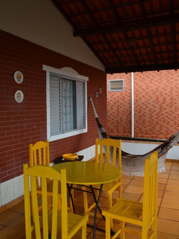Mesa e cadeiras de madeira amarelas numa varanda de tijolo, com rede ao fundo