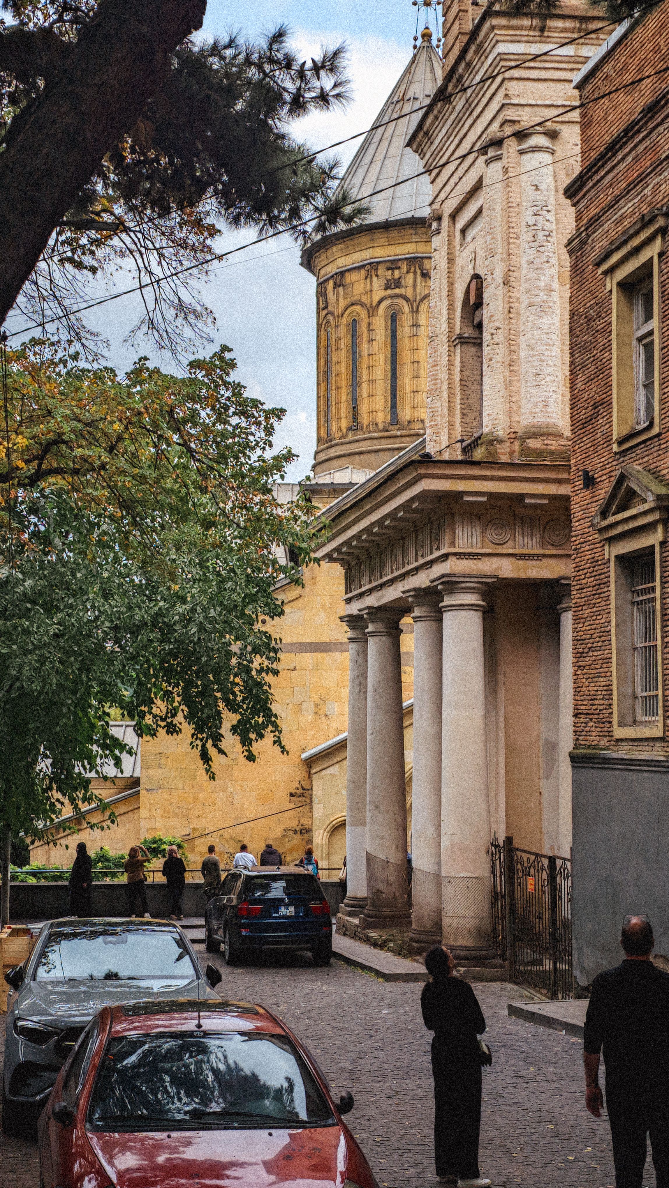 People walking on a historic street beside an ancient church in Tbilisi, Georgia.