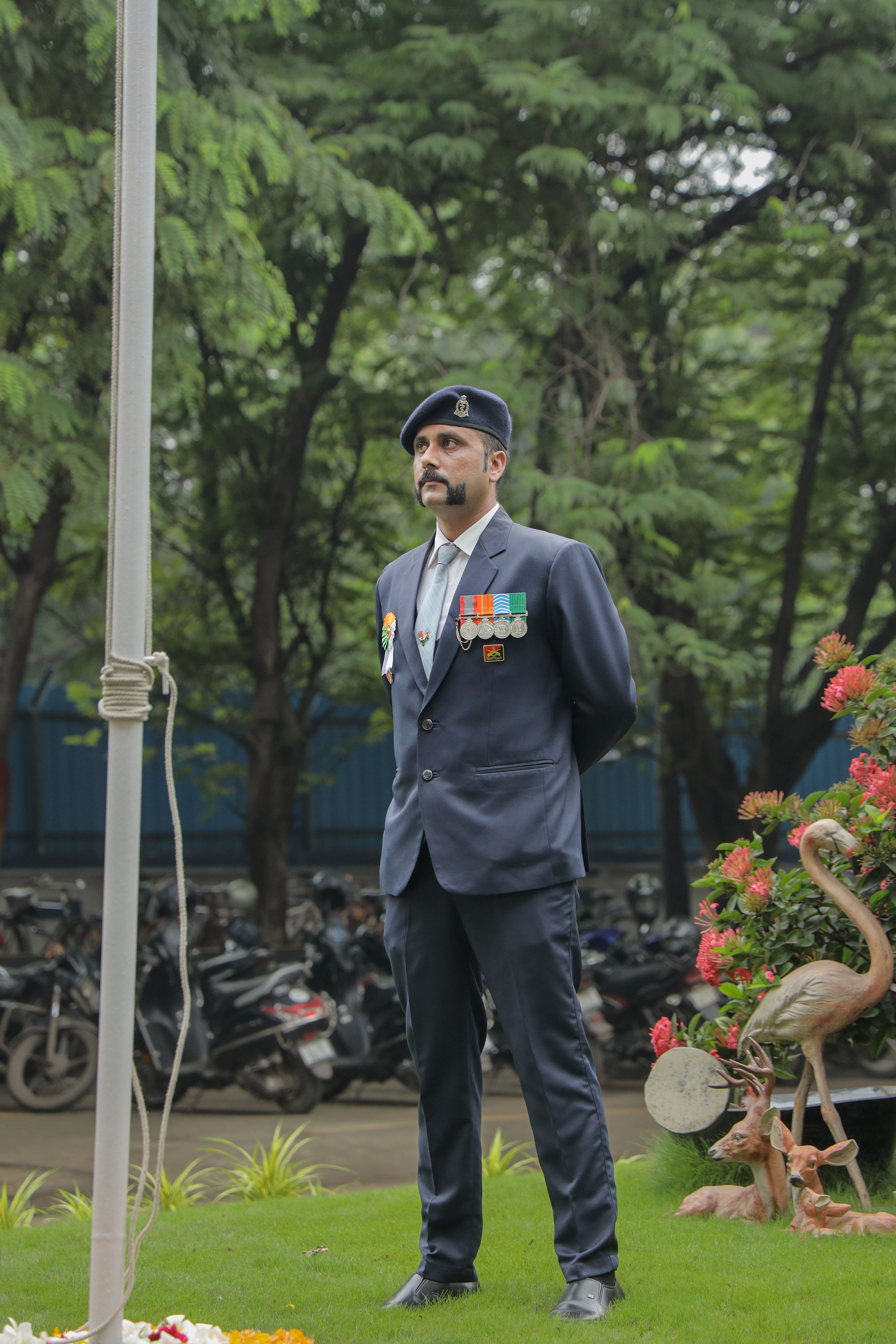 A proud military officer in uniform standing at a flag hoisting ceremony outdoors, surrounded by greenery.