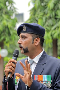A South Asian officer in a decorated uniform speaks into a microphone, outdoors.