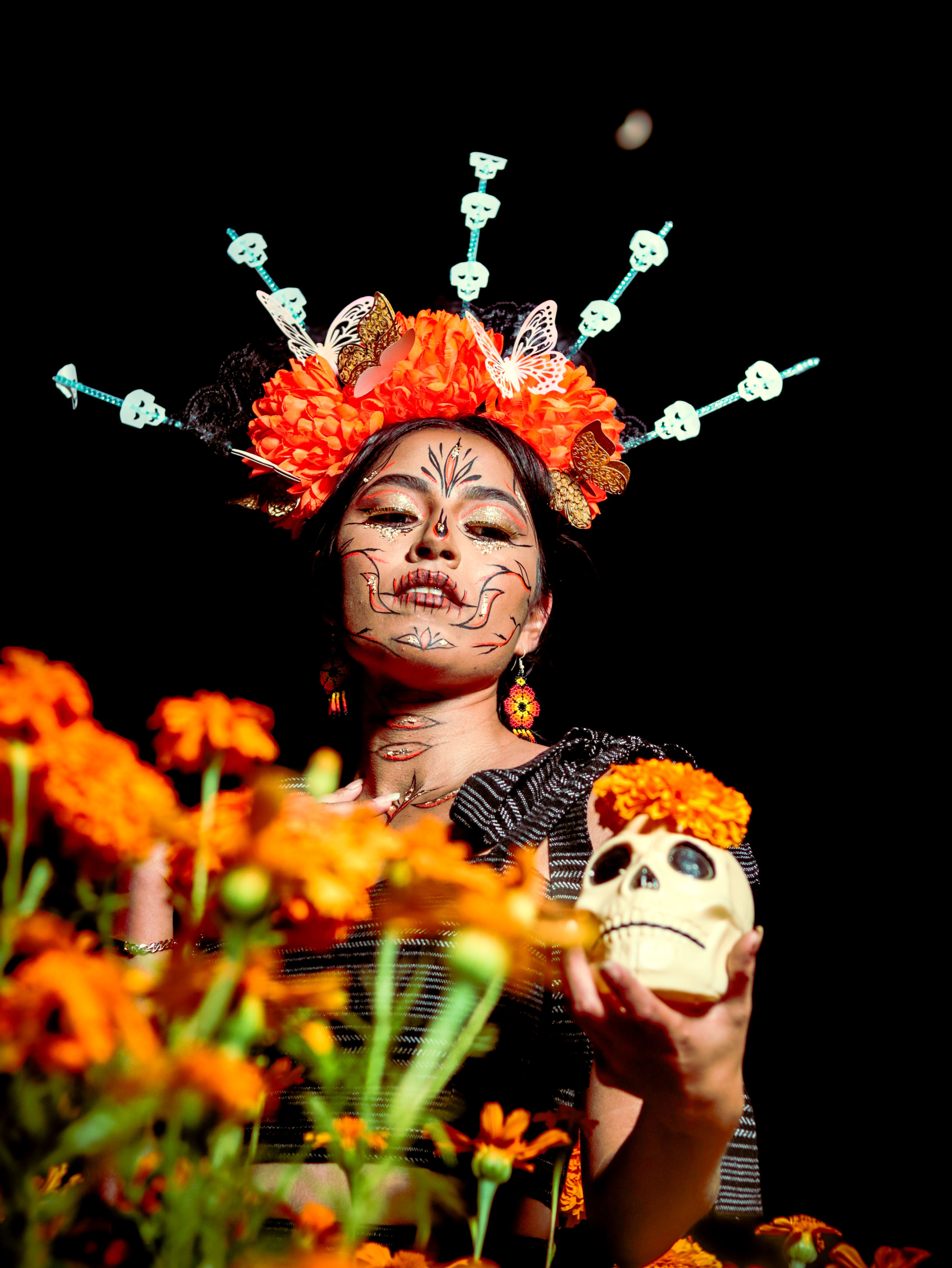 A woman in traditional Dia de los Muertos attire with marigolds and face paint, holding a skull.