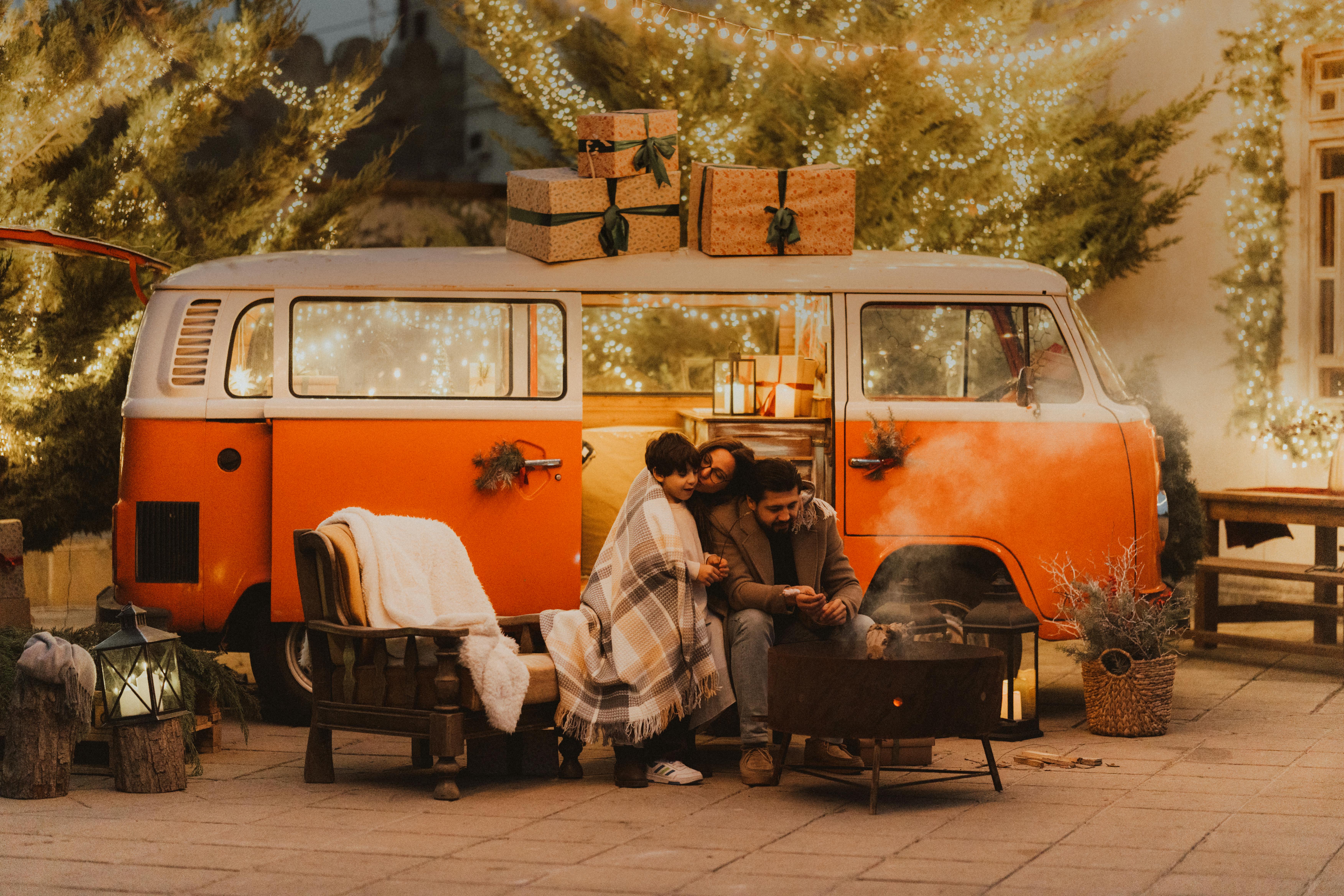 Family enjoys cozy evening by a decorated van with festive lights and gifts outdoors.