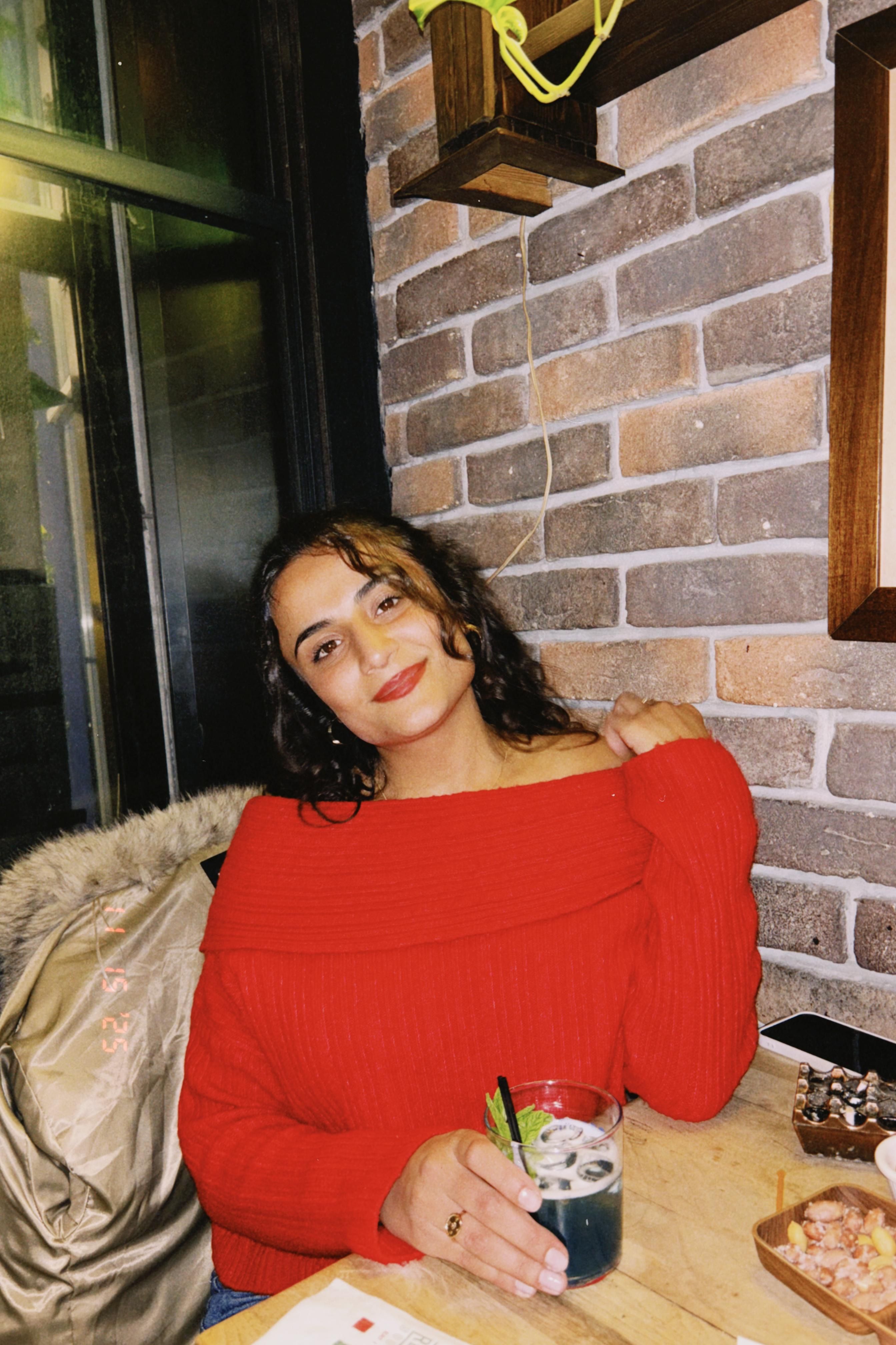 Young woman in a cozy red sweater enjoying a drink in a stylish indoor setting with brick walls.