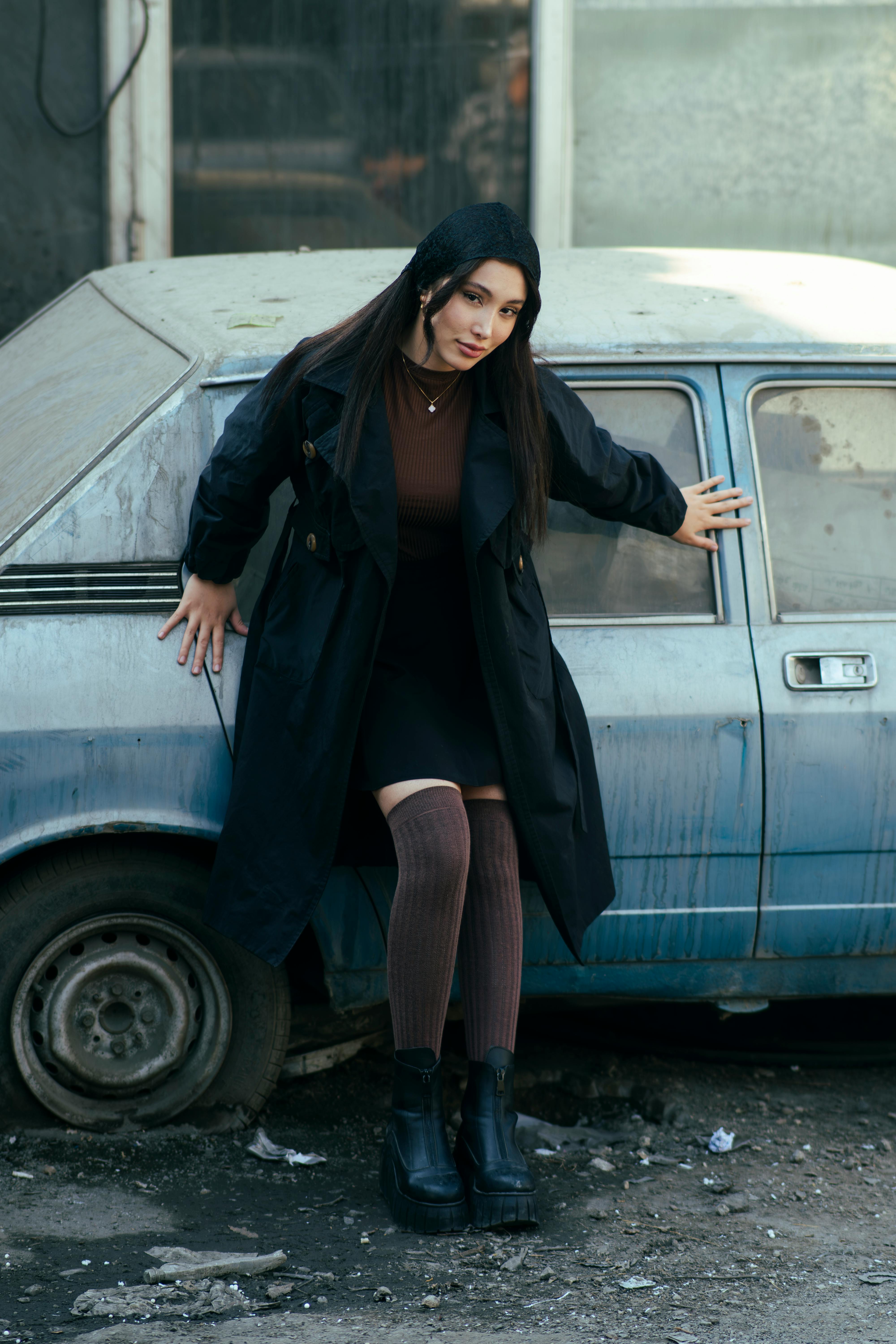 A fashionable woman in a black coat leans against a vintage car on an urban street in Tehran.