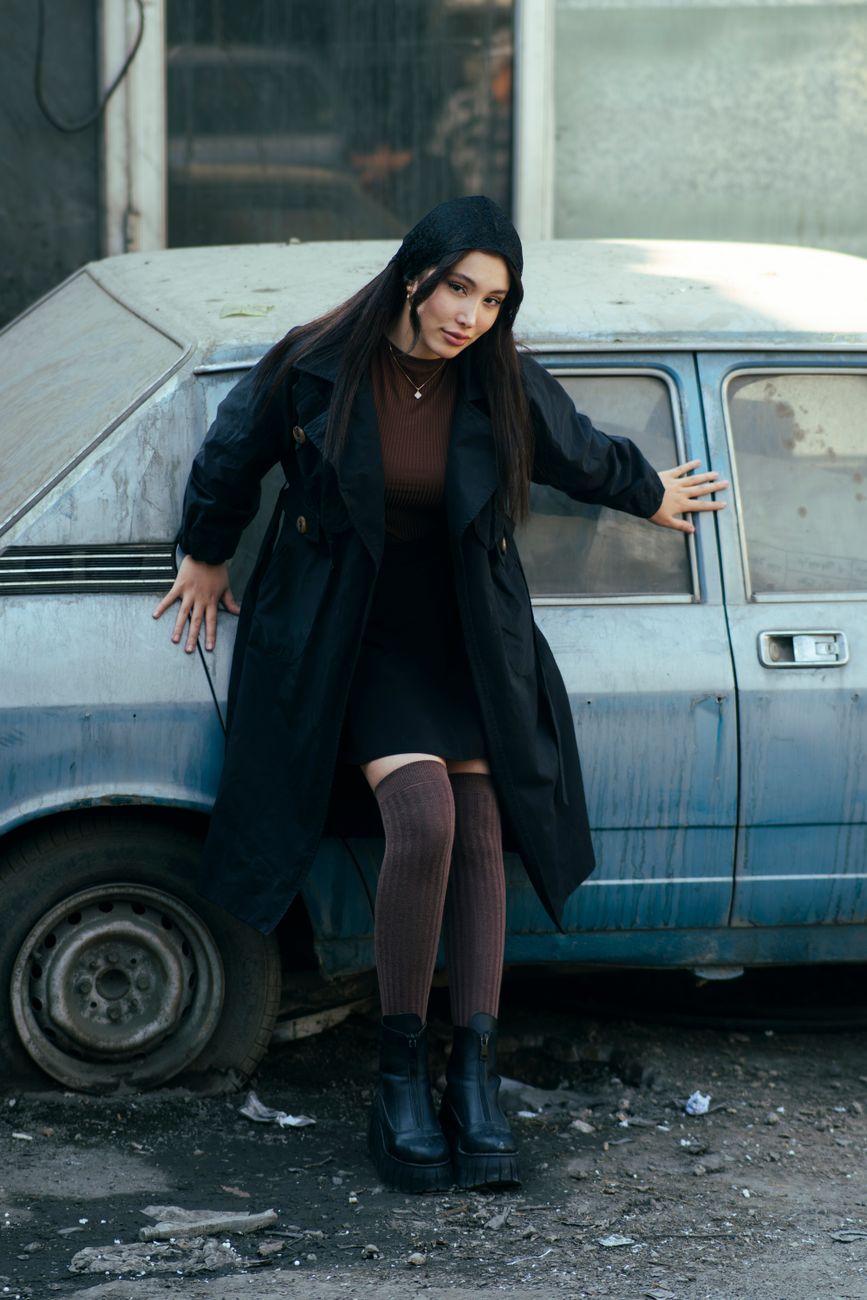 A fashionable woman in a black coat leans against a vintage car on an urban street in Tehran.