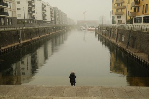 Free A lone figure stands by a foggy canal in Mainz, Germany, surrounded by urban architecture. Stock Photo