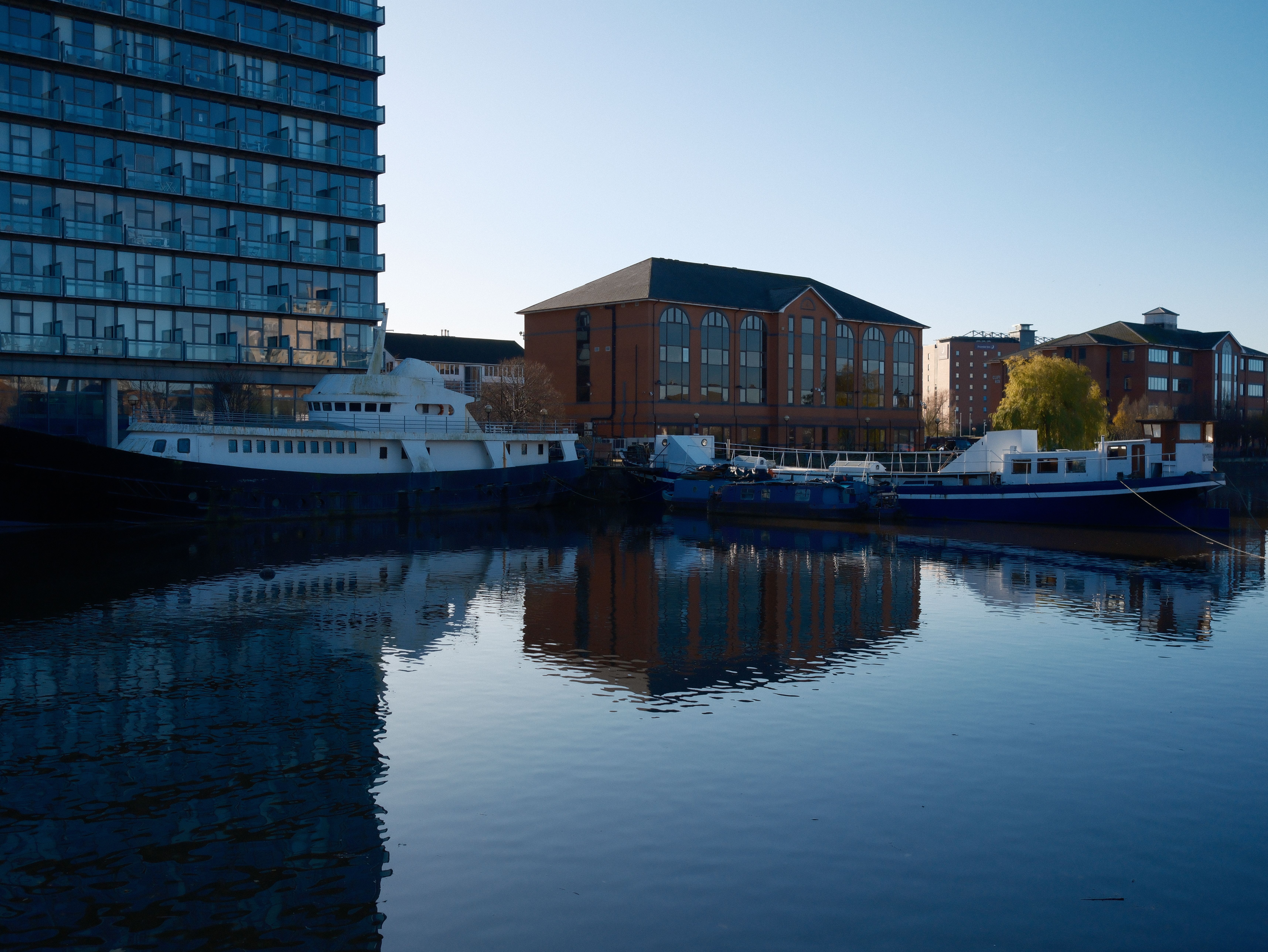 Boats docked next to modern and classic buildings reflecting on calm waters under a clear blue sky.