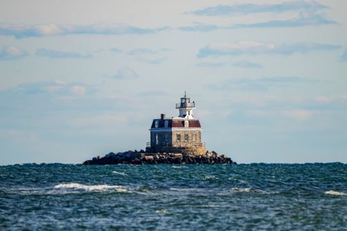 Free A solitary lighthouse standing on a rocky island surrounded by ocean waves under a blue sky. Stock Photo