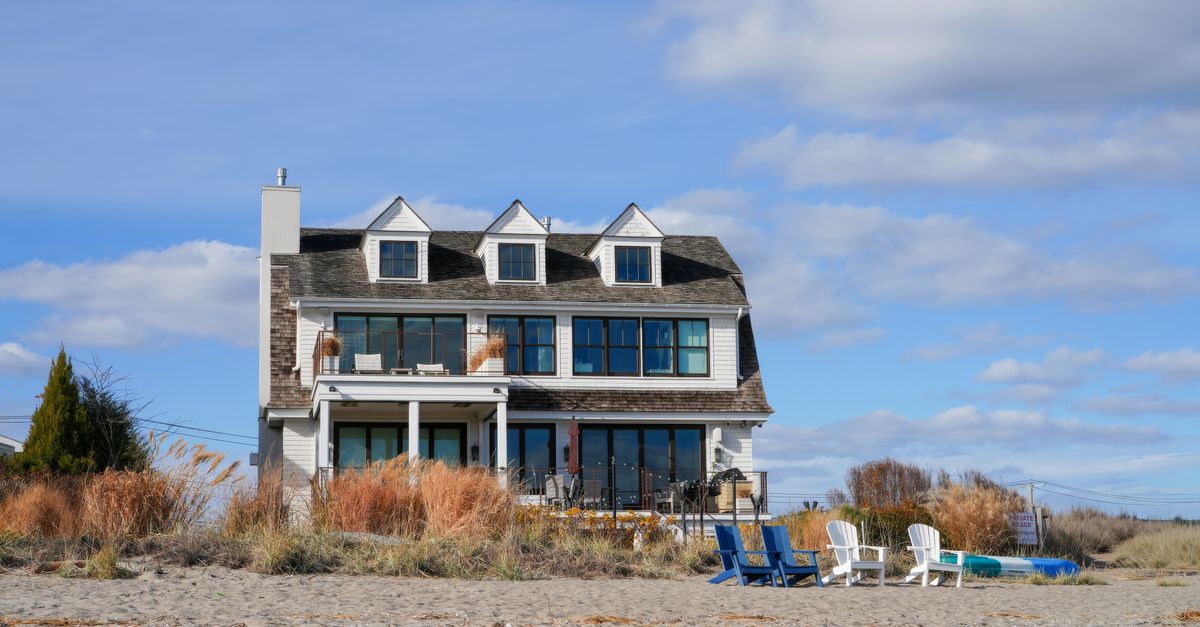 Cozy beachfront house with patio chairs surrounded by sand and grasses under blue sky.