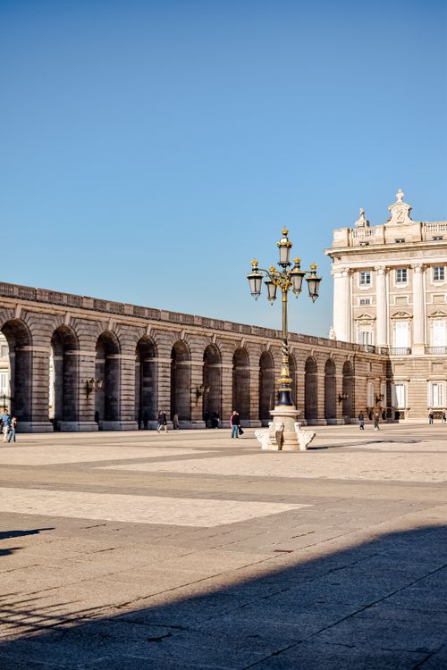 Free Elegant view of Royal Palace of Madrid courtyard with archways and lamp post under blue skies. Stock Photo