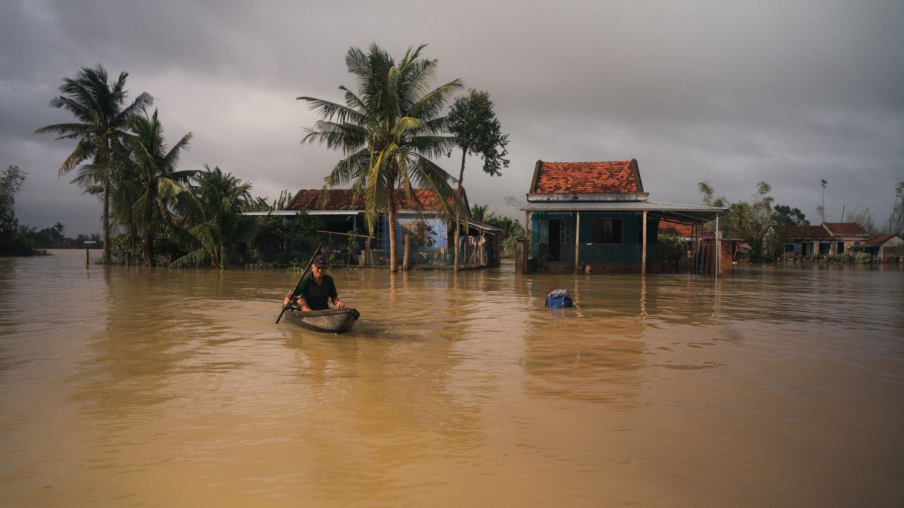 A man paddles a boat through a flooded village in Phú Yên, Vietnam, under cloudy skies.