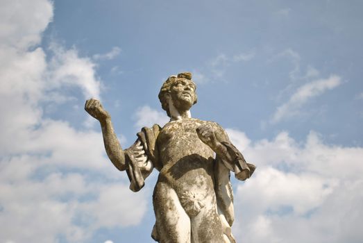 A weathered stone statue standing tall against a bright blue sky with clouds, showcasing classic artistry.