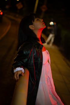 A young woman in a leather jacket and white dress relaxes under red city lights at night in Miraflores, Lima.