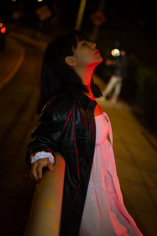 Free A young woman in a leather jacket and white dress relaxes under red city lights at night in Miraflores, Lima. Stock Photo
