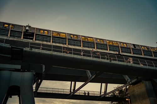 Free City train crosses an elevated bridge at sunset with people walking below. Urban transit scene. Stock Photo