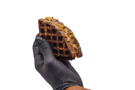 Close-up of a gloved hand holding a chocolate-covered waffle with nuts.