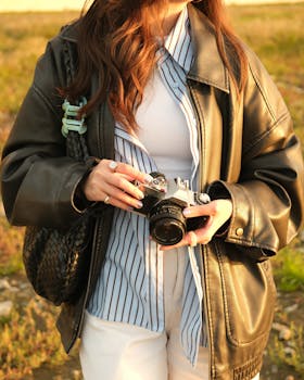 A woman in a leather jacket holds a camera in a sunlit outdoor setting.