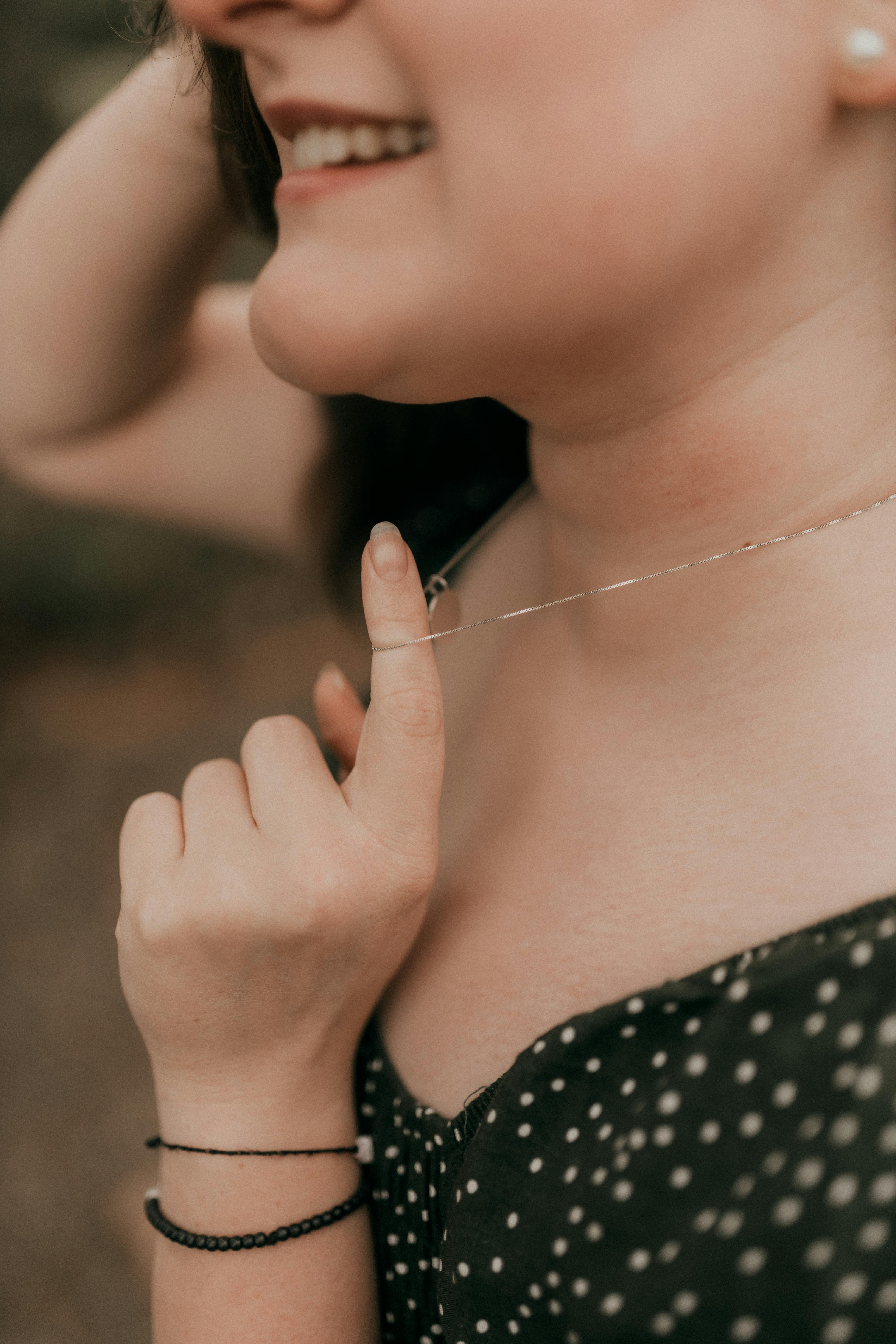 Gratis Gambar close-up seorang wanita tersenyum sambil menyentuh kalungnya dengan lembut, melambangkan keanggunan dan kegembiraan. Foto Stok