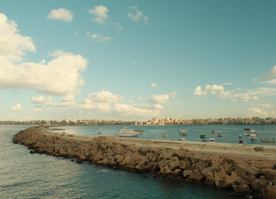 A picturesque view of a Mediterranean coastline with boats and distant city skyline under a partly cloudy sky.