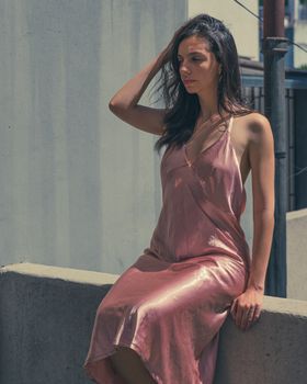 Elegant woman seated outdoors in sunlight, wearing a pink dress.