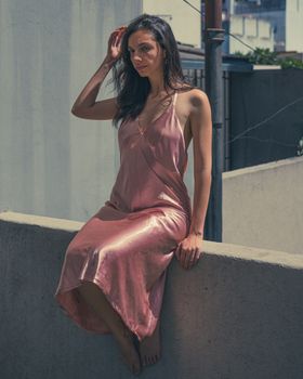A woman in a pink satin dress poses on a sunlit balcony, exuding elegance and style.