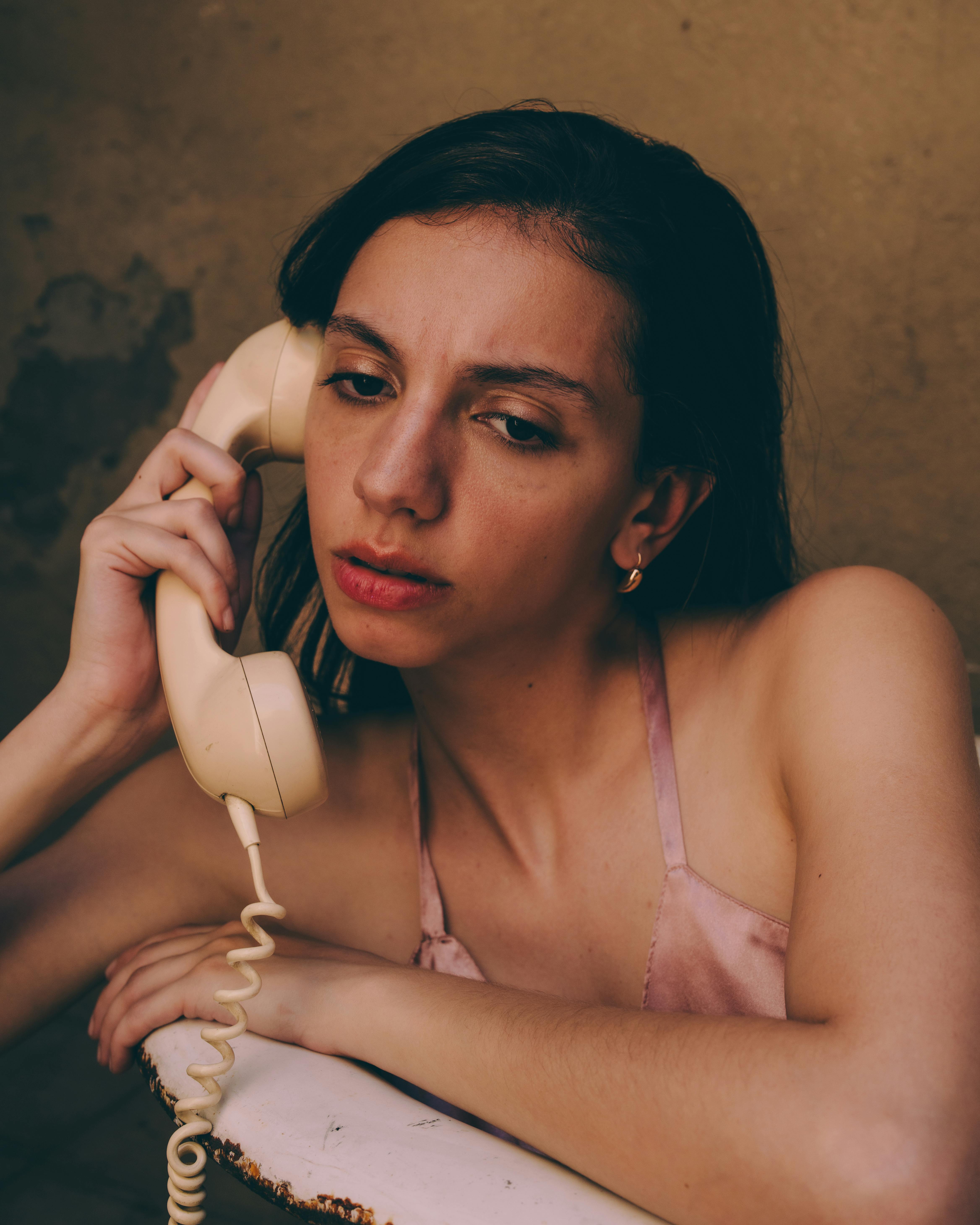 grátis Mulher pensativa falando ao telefone antigo dentro de casa, transmitindo nostalgia e emoção. Foto profissional