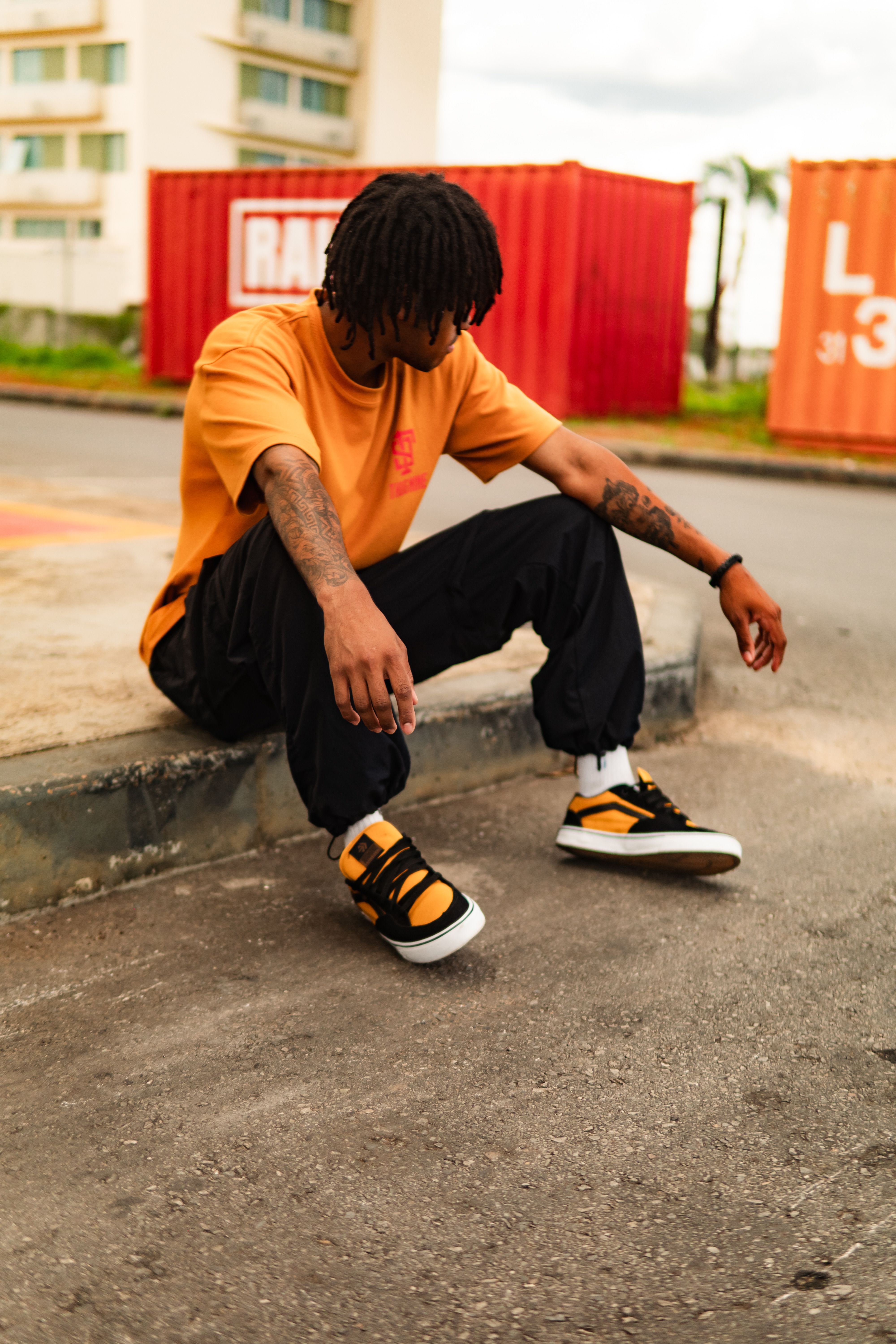 Young adult in casual streetwear sitting on curb with containers in background.