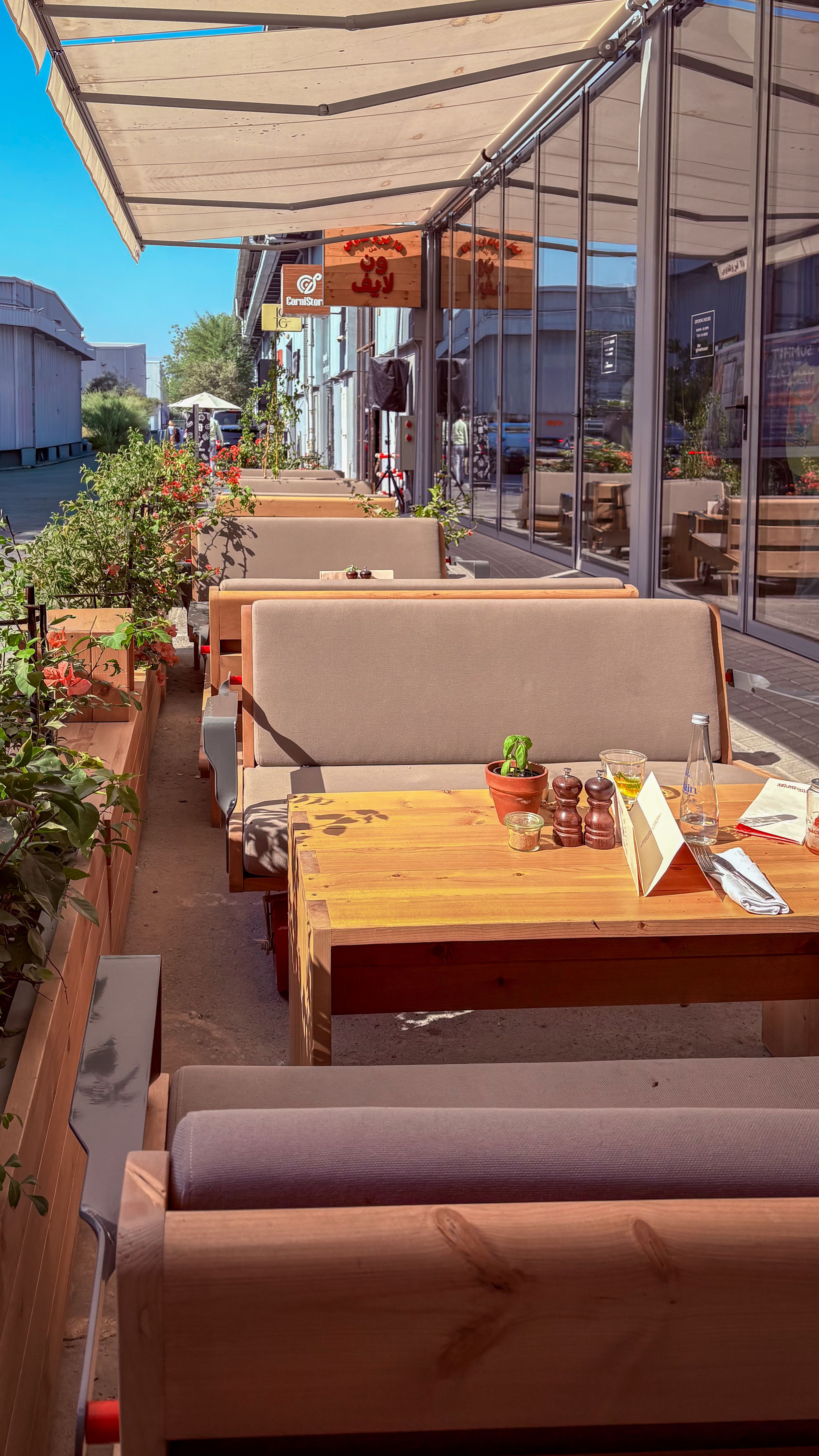 A bright outdoor cafe patio with modern wooden tables and cozy seating under awnings.
