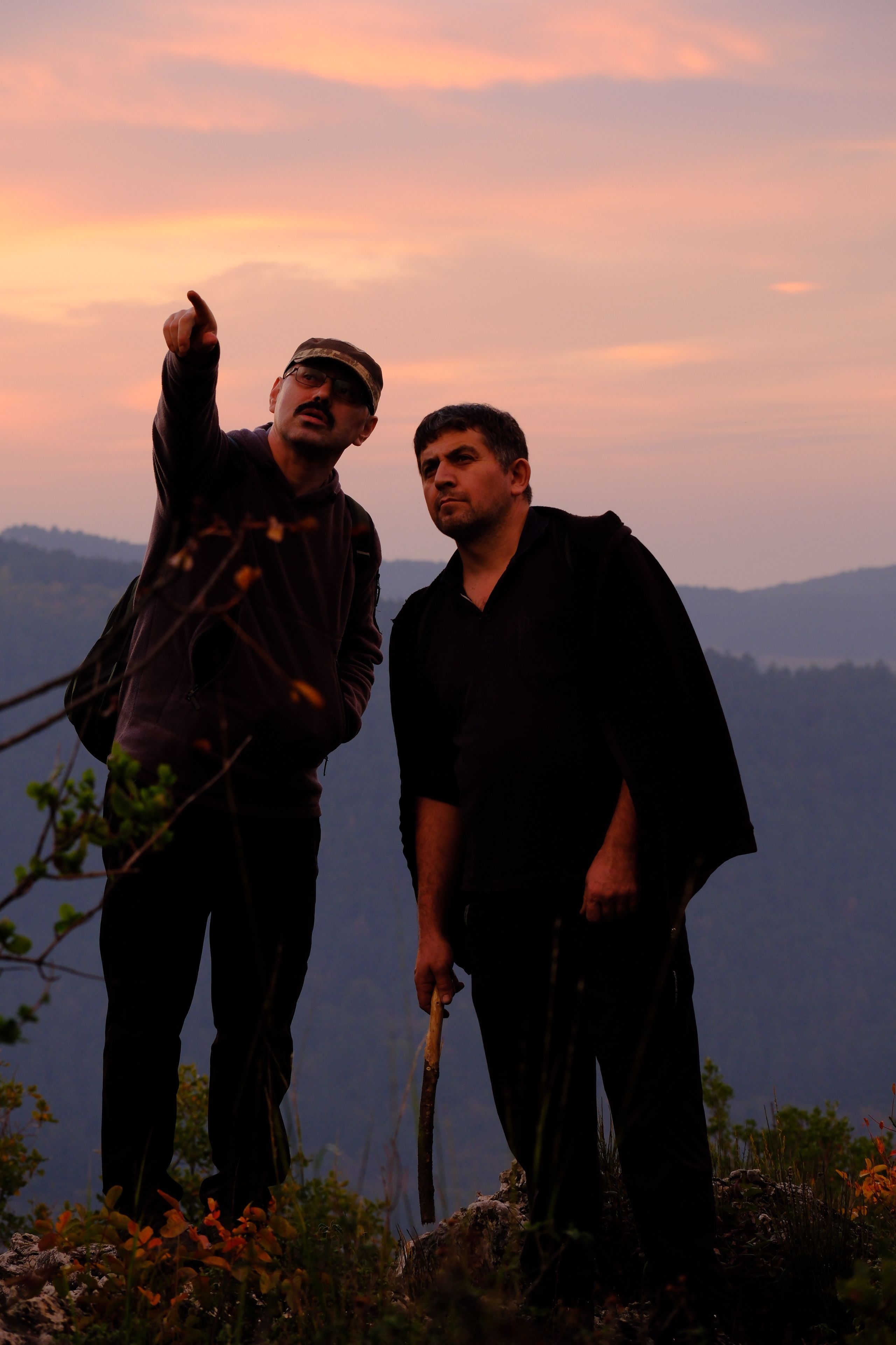 Two men standing on a hilltop at sunset, one pointing, against a mountain backdrop.