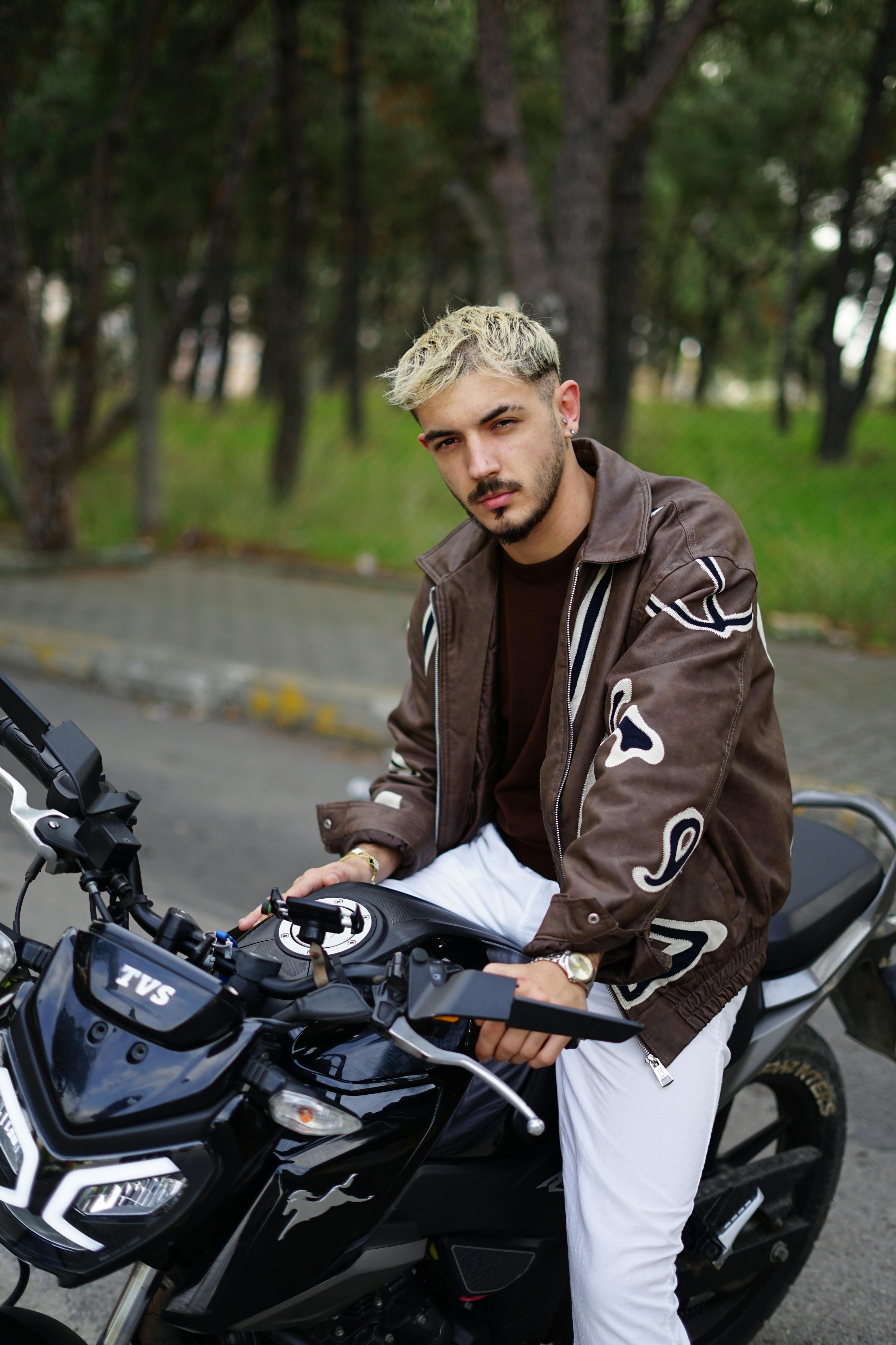 Stylish young man with blond hair on a motorcycle outdoors in casual attire.