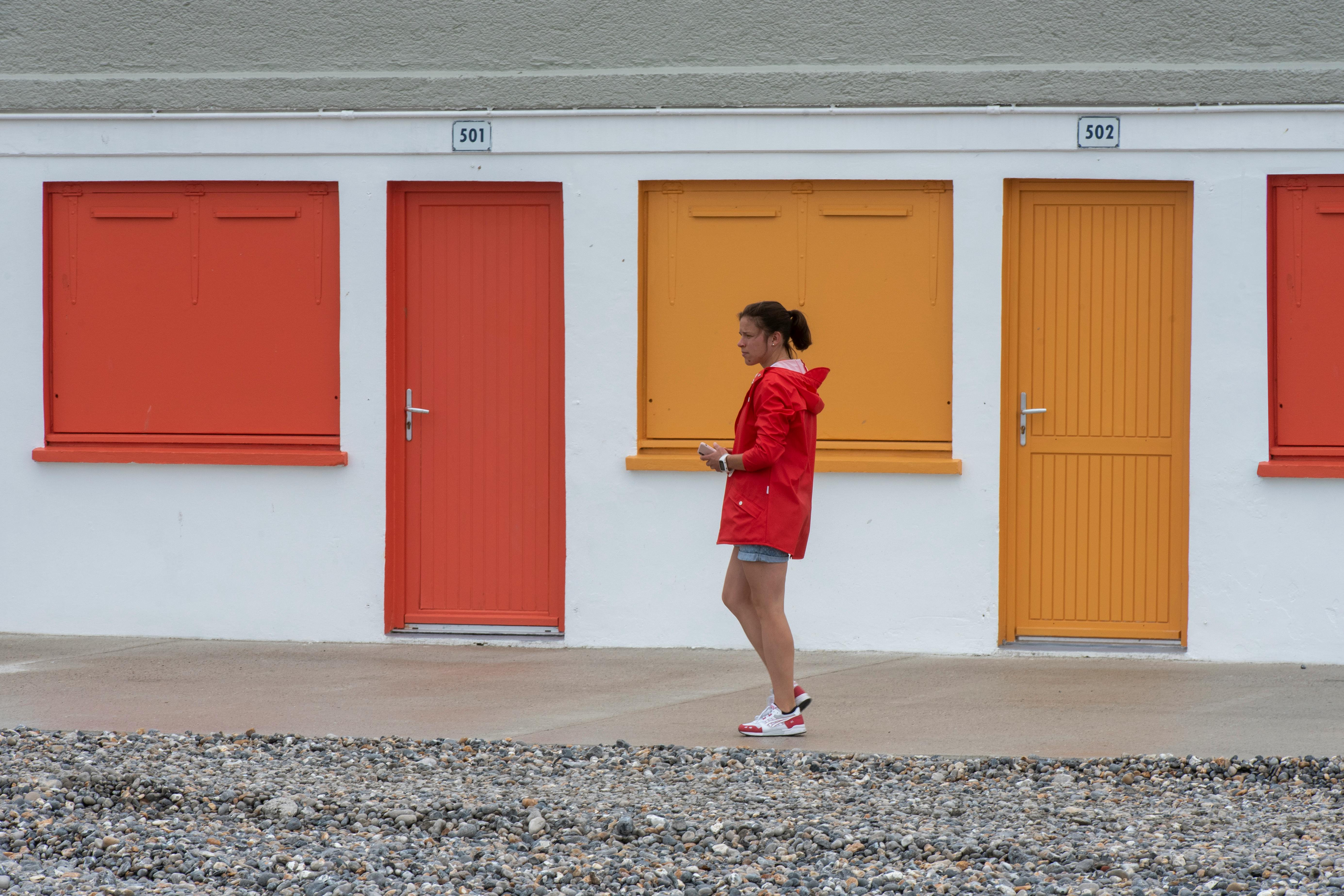 A woman walks past vibrant orange and red beach huts during a cool autumn day.