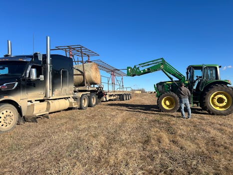 Tractor loads hay bales onto a truck in an open field under a clear blue sky.
