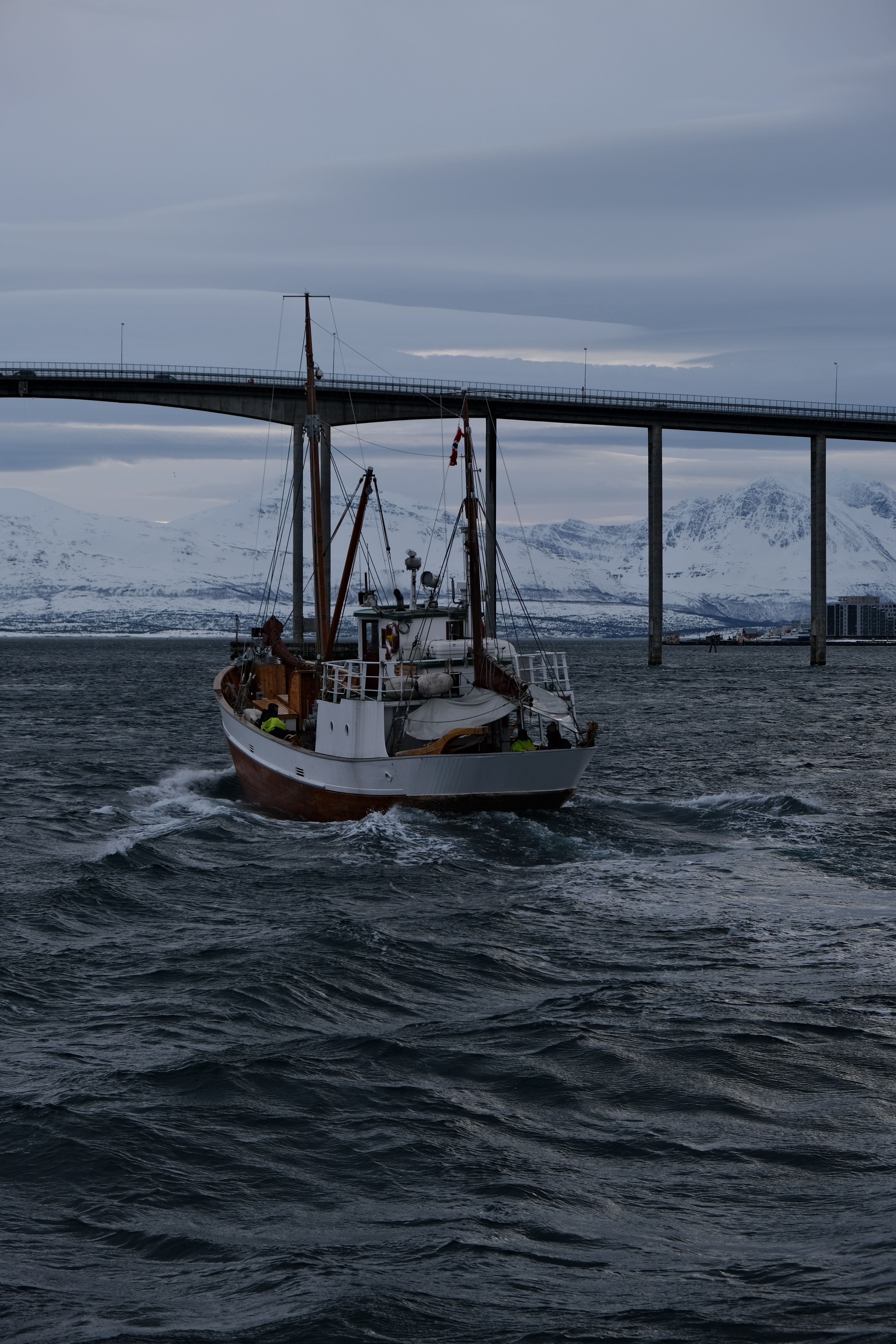 A fishing boat sails under a bridge with snowy mountains in the background.