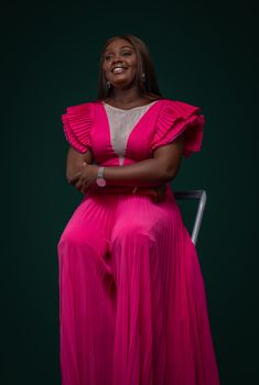 A joyful woman in a stylish pink dress seated on a chair in a studio setting.